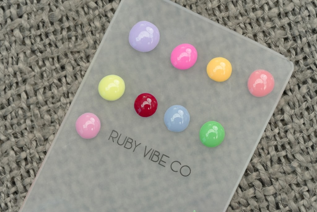 Swatches of ten different colorful, glossy gel nail polishes dotted on a clear Ruby Vibe Co palette resting on a gray textured surface.