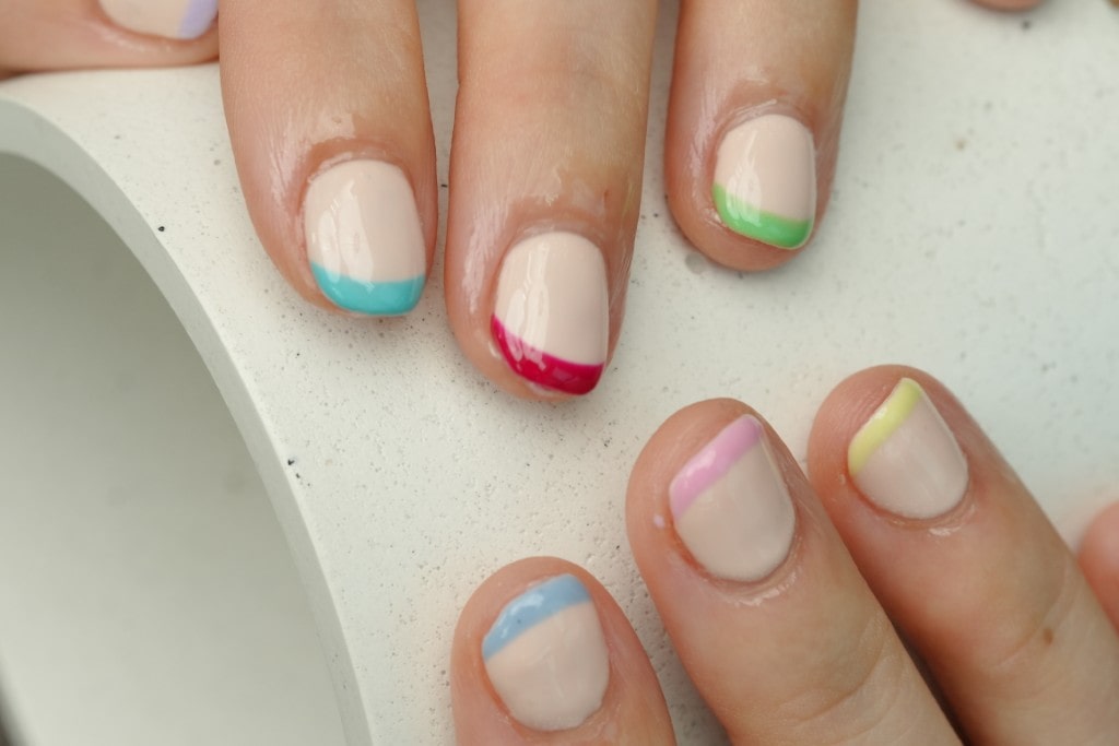  A close-up of a minimalist manicure on short nails, featuring a nude base with a different bright color painted as a thin, angled tip on each finger.
