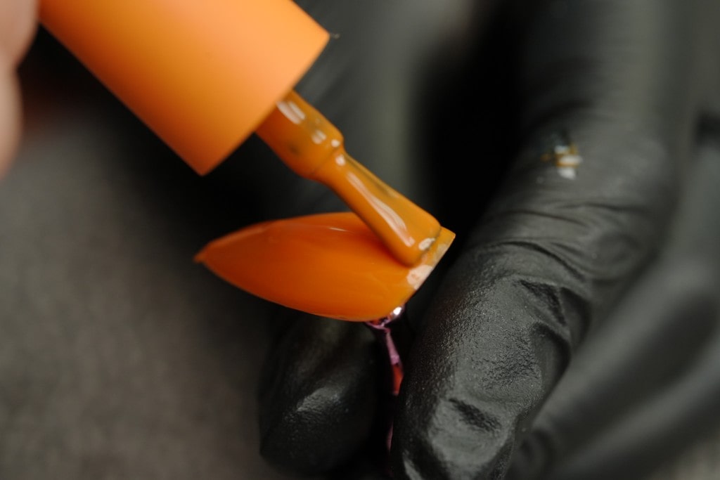 A macro shot of a bright orange nail polish being applied with a brush to a long, pointed nail tip held by a black-gloved hand.