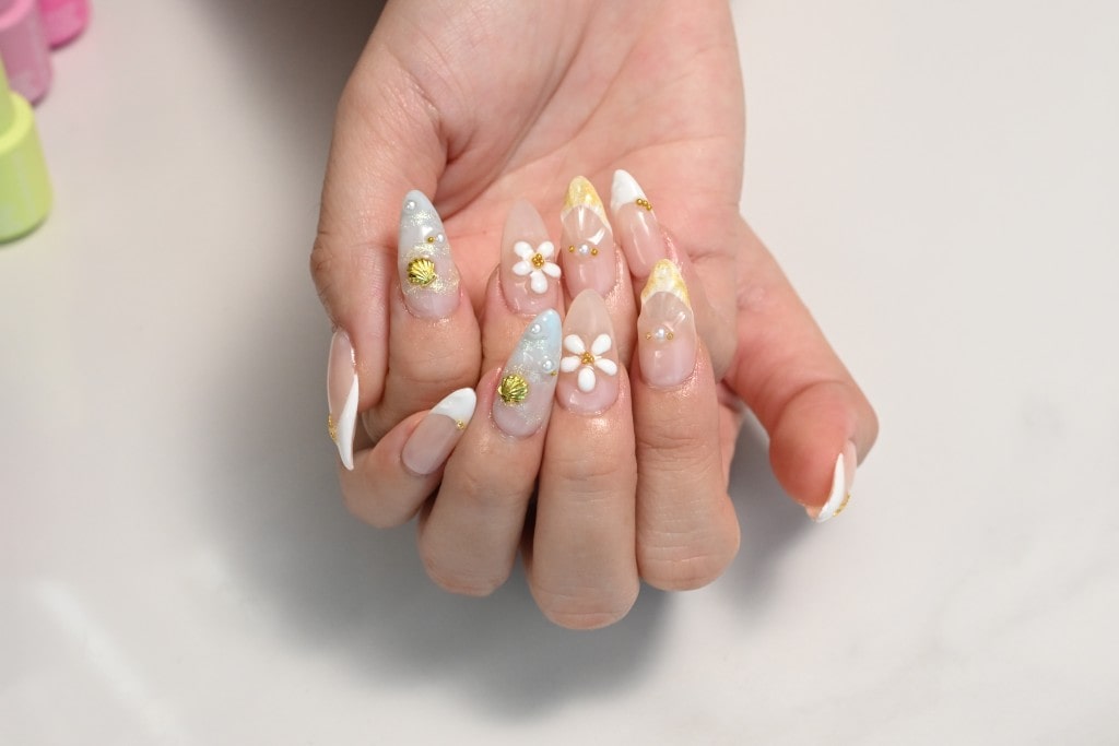  A close-up of a hand with an elaborate, glam manicure featuring long almond-shaped nails with 3D flowers and gold embellishments.