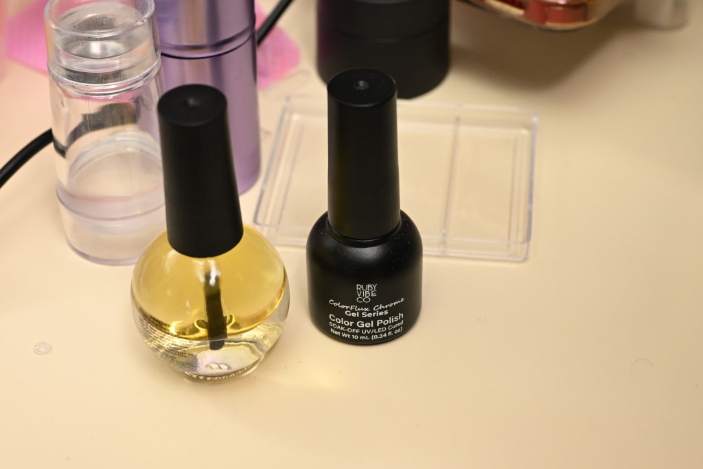 A bottle of cuticle oil next to a black bottle of Ruby Vibe Co. ColorFlux Chrome Color Gel Polish.