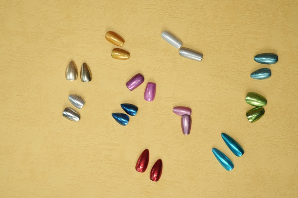 An overhead view of colorful, metallic press-on nails in various shapes and shades scattered on a tan background.
